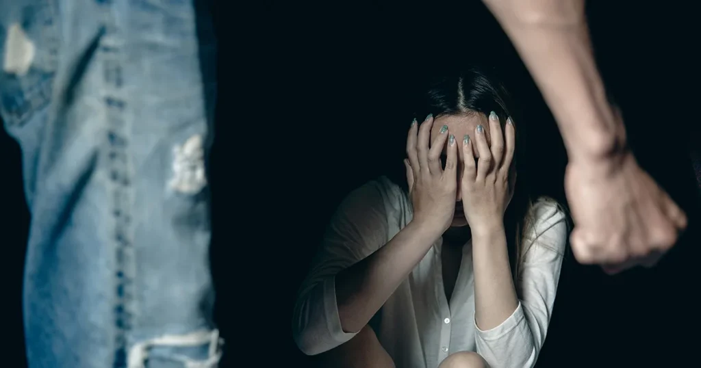 Distressed woman hides her face with both hands as a person stands nearby with a raised fist.