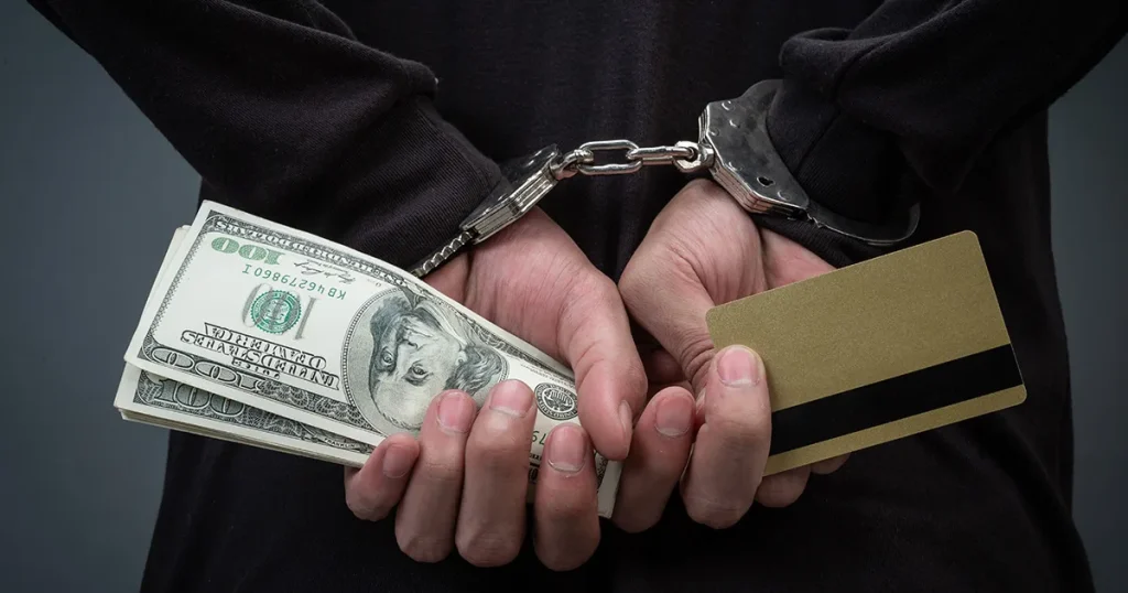 Close-up of handcuffed hands behind a person's back, clutching US currency notes and a plastic credit card, illustrating financial arrest.