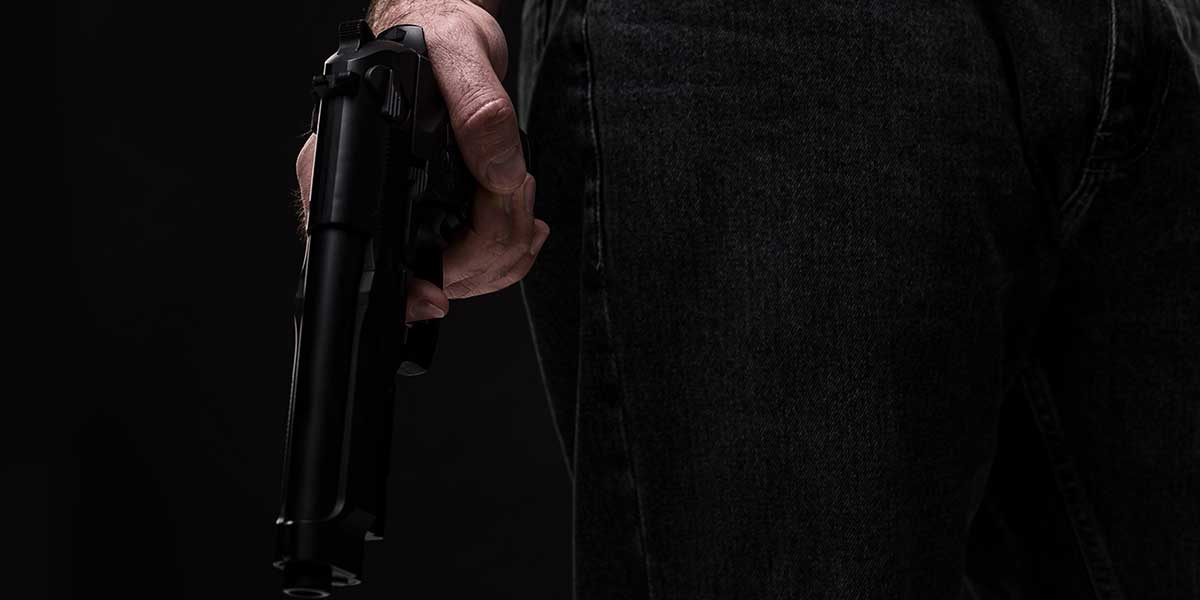 A close-up view of a person holding a black handgun at their side against a dark background in shadow.